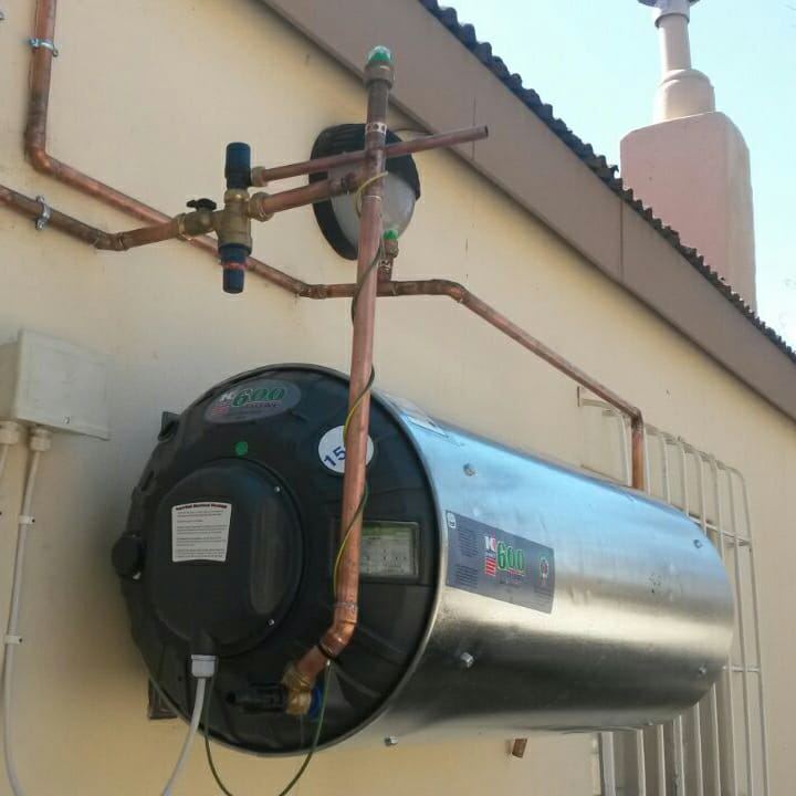 A stainless steel geyser mounted on an outside wall with copper pipes and valves